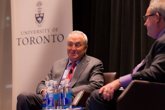 Lorne Michaels smiles and talks, sitting in a chair on a stage. A sign behind him reads, University of Toronto.
