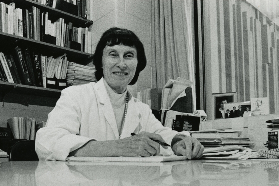 Marian Packham smiles as she sits at an office desk, wearing a lab coat.