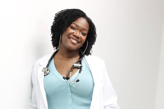 Onye Nnorom leans against a wall, smiling, wearing lab coat and stethoscope.