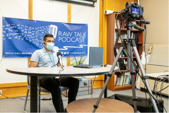 Yagnesh Ladumor, wearing a mask, sits at a table with a large microphone, in front of a banner that reads Raw Talk Podcast.