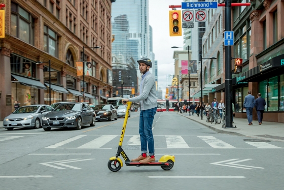 A young man wearing a helmet stands on an e-scooter as he crosses College Street in downtown Toronto.