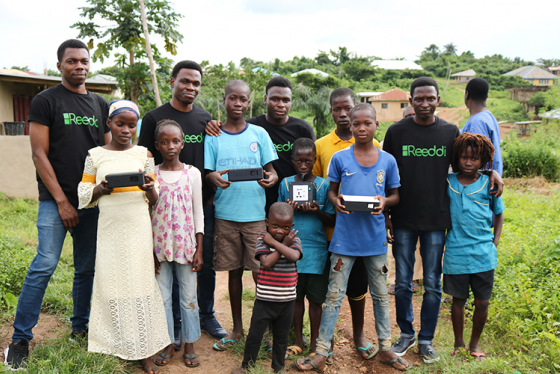 Four men wearing Reedi T-shirts pose with a group of children holding Reedi devices in a village among trees.