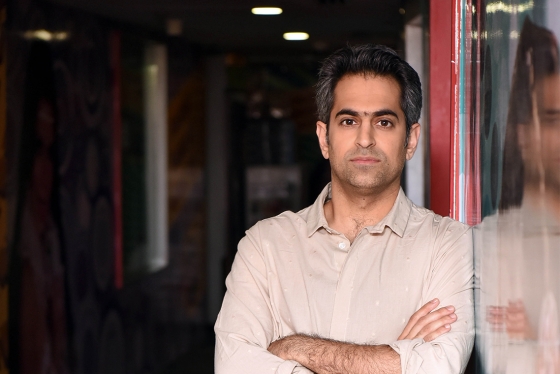 Richie Mehta looks serious as he leans on a glass wall indoors with arms folded.