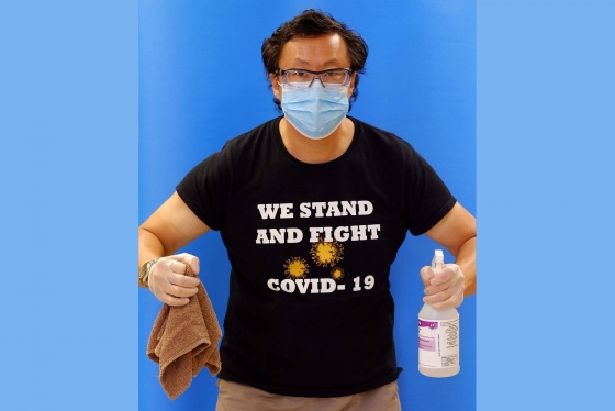 Stephen Lew wears a mask and a T-shirt reading We Stand and Fight COVID-19. He holds cleaning supplies in his gloved hands.