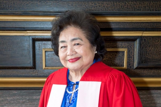 Setsuko Thurlow smiles, weating academic robes and standing against a wood-panelled wall.