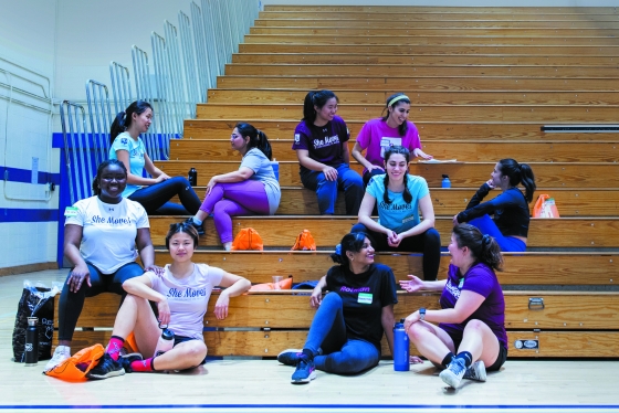 A diverse group of smiling students chat on the bleachers in a gym, wearing She Moves t-shirts.