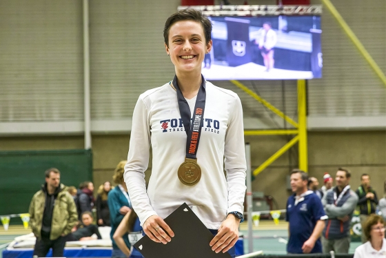 Gabriela DeBues-Stafford smiles, standing in a gym wearing a U of T shirt and a gold Track and Field 2017 medal.