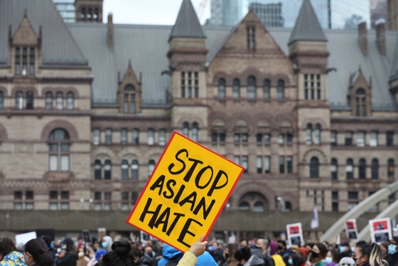 A handlettered sign reading Stop Asian Hate is held high above a crowd in front of Toronto's Old City Hall.