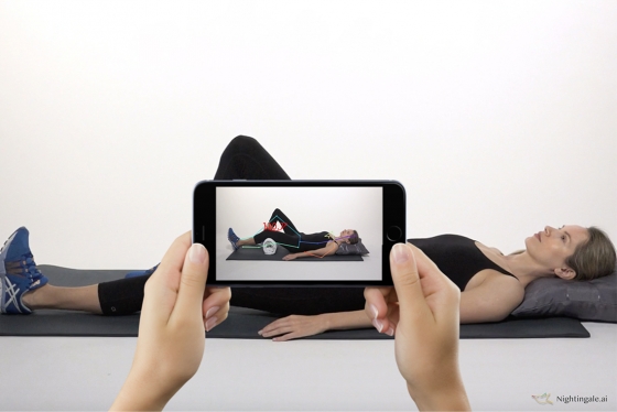 Two hands hold a phone to film a woman on an exercise mat. Lines overlay her body on screen, showing the angles of her limbs.