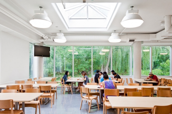 Students eat and chat at wooden tables in a room with one glass wall overlooking a lush garden.