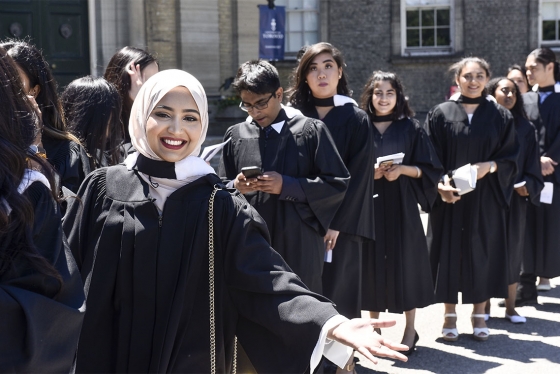 A woman smiles and gestures while standing in a line of diverse students in academic robes waiting to graduate.