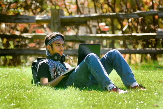 A young man sits alone outdoors on the grass, looking at his laptop screen.