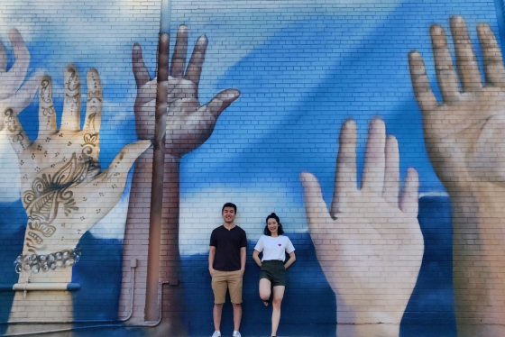 Weiwei Li and Catherine Chan smile as they pose beside a wall mural featuring several giant raised hands.