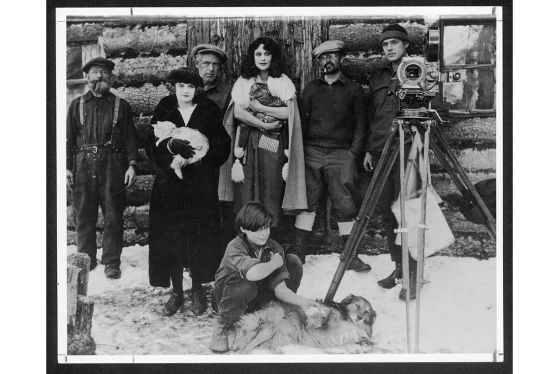 In 1923, director Nell Shipman stands in snow in front of a log cabin, with a film camera and several crew and cast members.