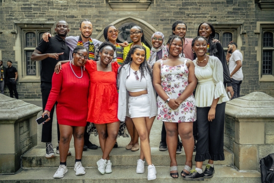 A group of 12 smiling Black graduates and their friends and family stand outside of Hart House.