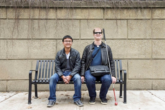 Rylan Vroom, who uses a white cane, and Bin Liu, who does not, smile as they sit together on a bench.