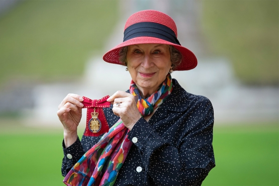 Margaret Atwood smiles and holds up a medal topped with a crown and mounted on a ribbon tied in a bow.
