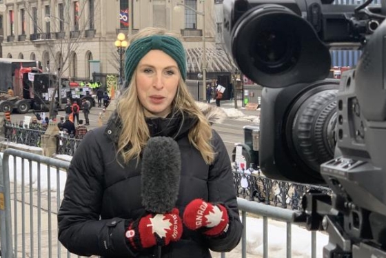 Annie Bergeron-Oliver holds a microphone and speaks to a camera in Ottawa, with protesting trucks behind her.