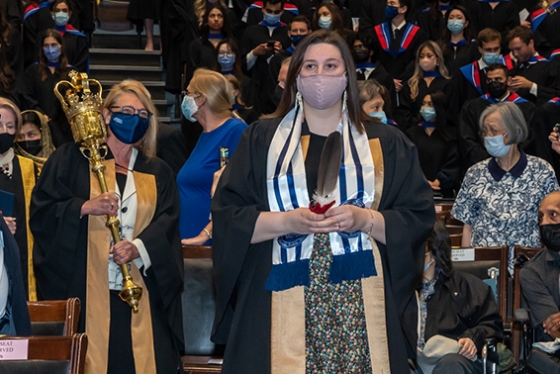 The audience in Convocation Hall stands as Lindsey Fechtig walks down an aisle holding an eagle feather.