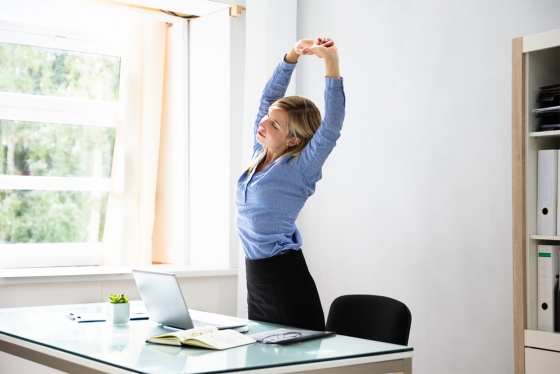A woman stands beside a desk, stretching up with her arms above her head.