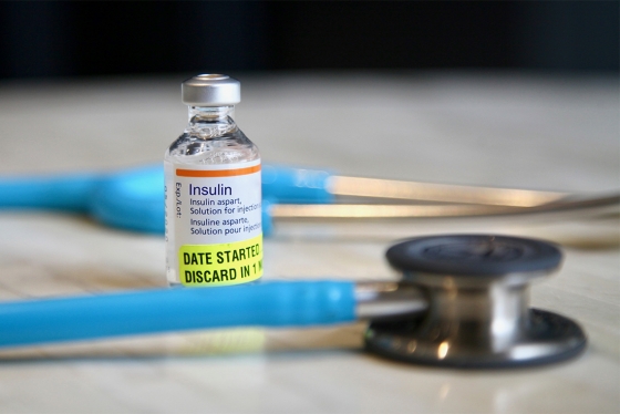 A vial labelled Insulin sits on a table, inside the curve of a doctor's stethoscope.
