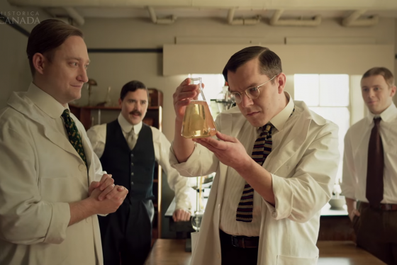 In a screenshot from the Heritage Minute on the discovery of insulin, 4 scientists in 1921 gaze at a beaker of clear liquid.
