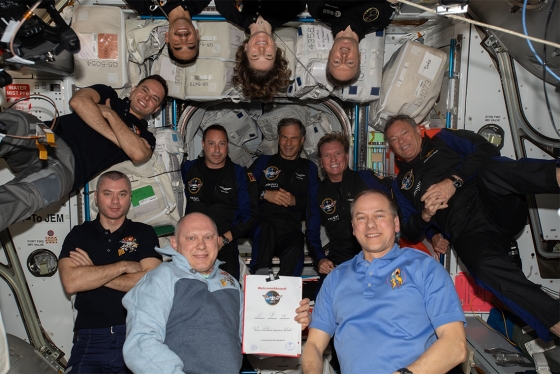 11 people pose together, floating in the air in a space station.
