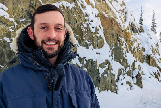 Luke Spooner smiles, as he stands in front of a snow-covered cliff, in a spruce forest.