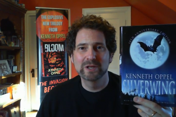 Kenneth Oppel smiles and holds up his book Silverwing in this still from a video.