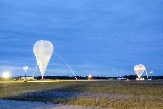 Two giant balloons, anchored in a field at dusk, gradually swell with hot air. The gondolas beneath are too small to see.