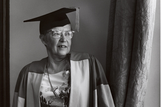 In a historical photograph, Elsie McGill stands by a window, wearing her academic hat and robes.