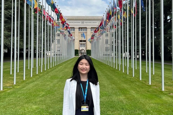Yifan Zhou smiling. standing next to the rows of flags outside the United Nations building in Geneva.