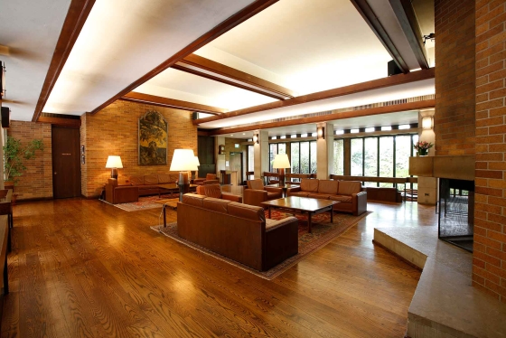 Image of Massey College's Common Room. There is brown furniture placed in the room to invite conversation. There are tables, lamps and a fireplace with large windows. Brick walls and tall white ceiling.
