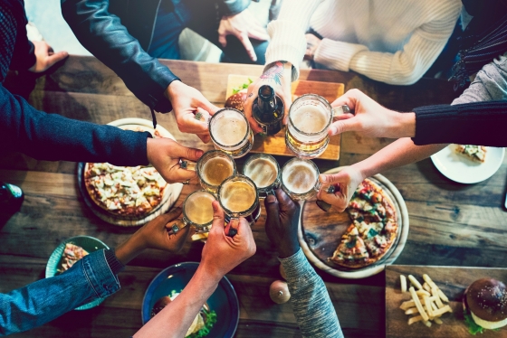A group of friend cheersing glasses over a wood bar table full of snacks.