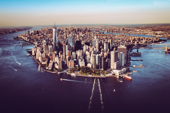 An aerial photo of Manhattan, New York City. You can see the iconic skyscrapers, the Hudson and East Rivers, and landmarks such as the One World Trade Center in the foreground and the Brooklyn and Manhattan Bridges in the distance.