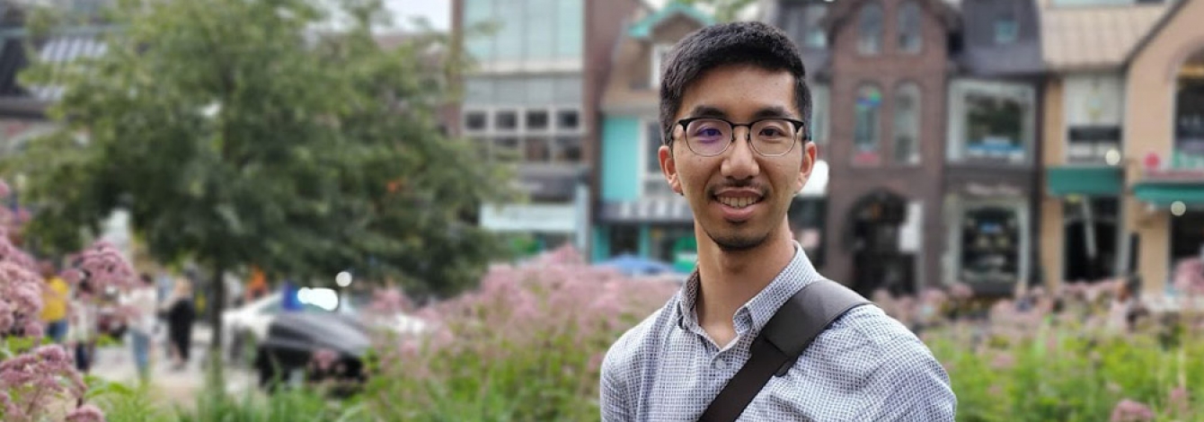 Lyndon Chan smiles while standing in a park beside a shopping street.