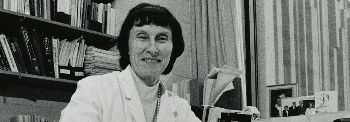 Marian Packham smiles as she sits at an office desk, wearing a lab coat.