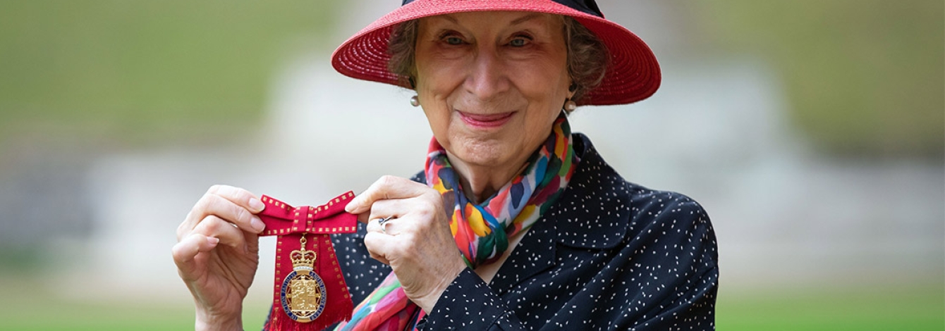 Margaret Atwood smiles and holds up a medal topped with a crown and mounted on a ribbon tied in a bow.