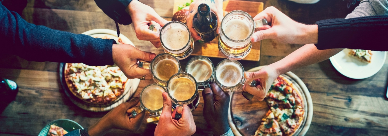 A group of friend cheersing glasses over a wood bar table full of snacks.