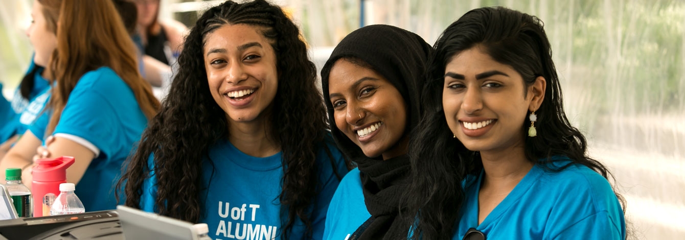Three women wearing Alumni Reunion T-shirts smile from behind a registration desk.