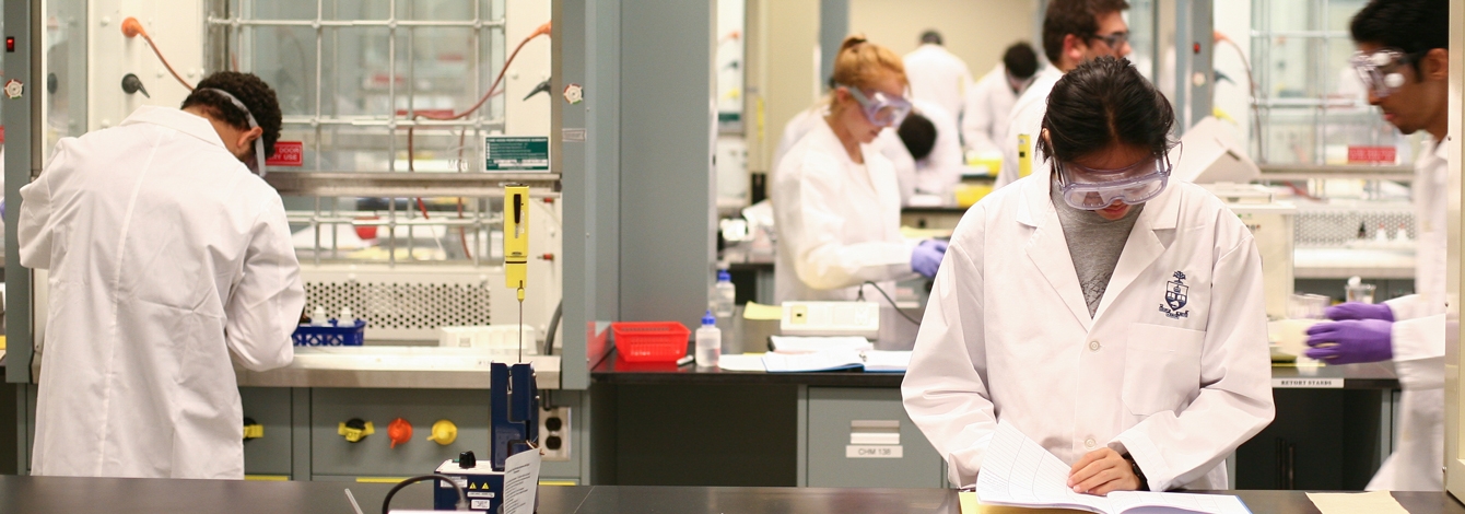 A diverse group of people in lab coats and safety goggles work intently the beakers, books and equipment in a lab at U of T's Department of Chemistry.