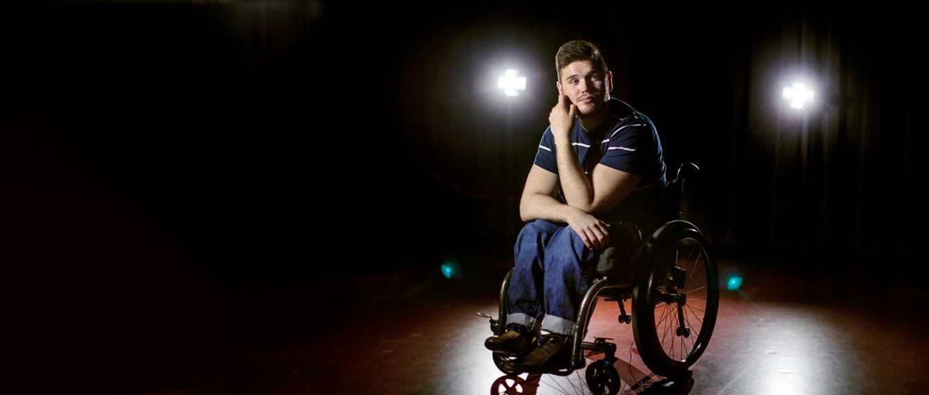 George Alevizos sits in his wheelchair, looking thoughtful, in the spotlight on a bare stage.