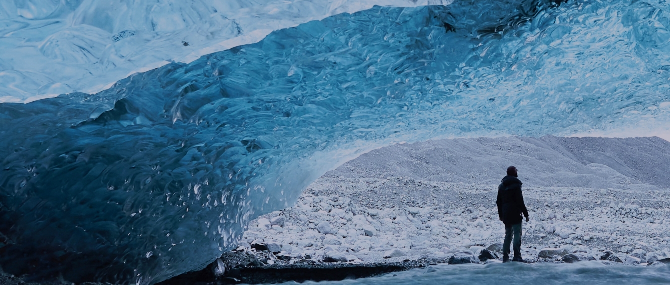 Man stands underneath an ice bridge looking at a mountain in the distance.