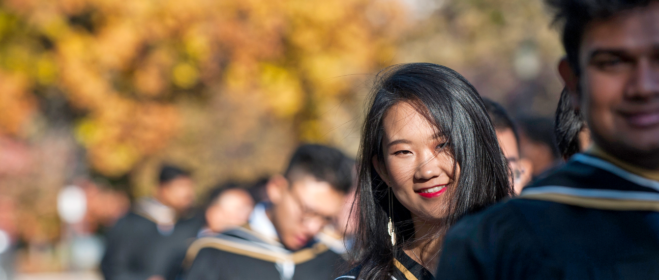 Young alum, smiling in her Convocation robes amidst the fall foliage