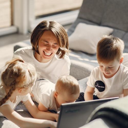 Woman sits on floor next to sofa with three kids looking at a laptop