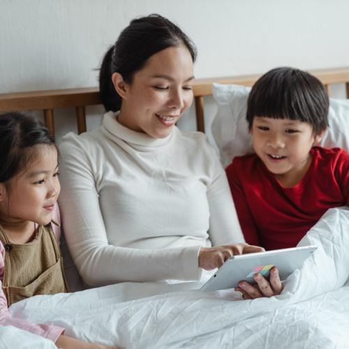 Mother sits with son and daughter on bed looking at tablet