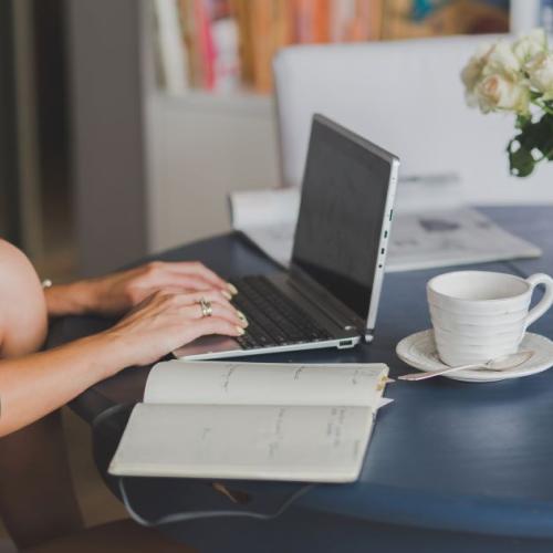 Woman sits at table with laptop, cup of coffee, open book, magazine and vase of flowers