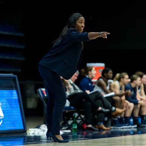 Black woman in navy blue suit stands on edge of basketball court pointing 