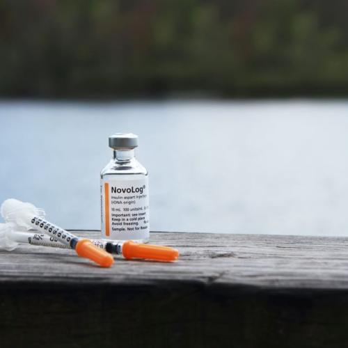 Vial of insulin sits with 2 needles on a wooden table