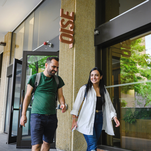Man in a green shirt and woman in a white button down, both wearing backpacks, walking outside of the OISE building.